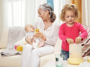<p>Granny holding the baby, sister is looking at the bottlewarmer </p>